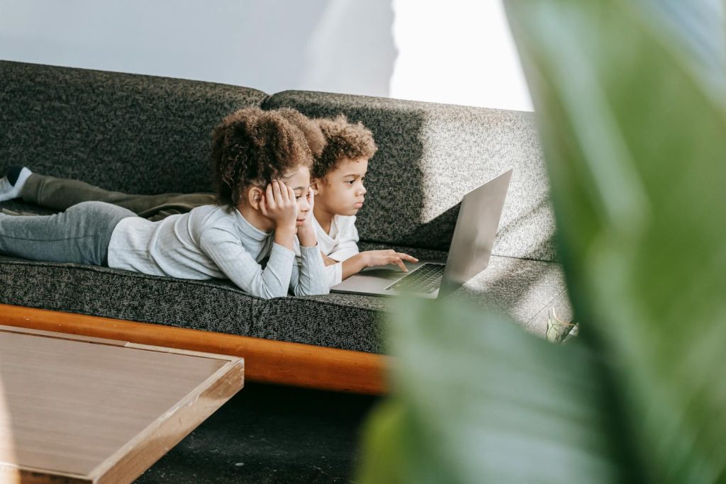 Side view of focused African American siblings surfing modern netbook while lying on comfortable sofa in room with wooden table and green leaves